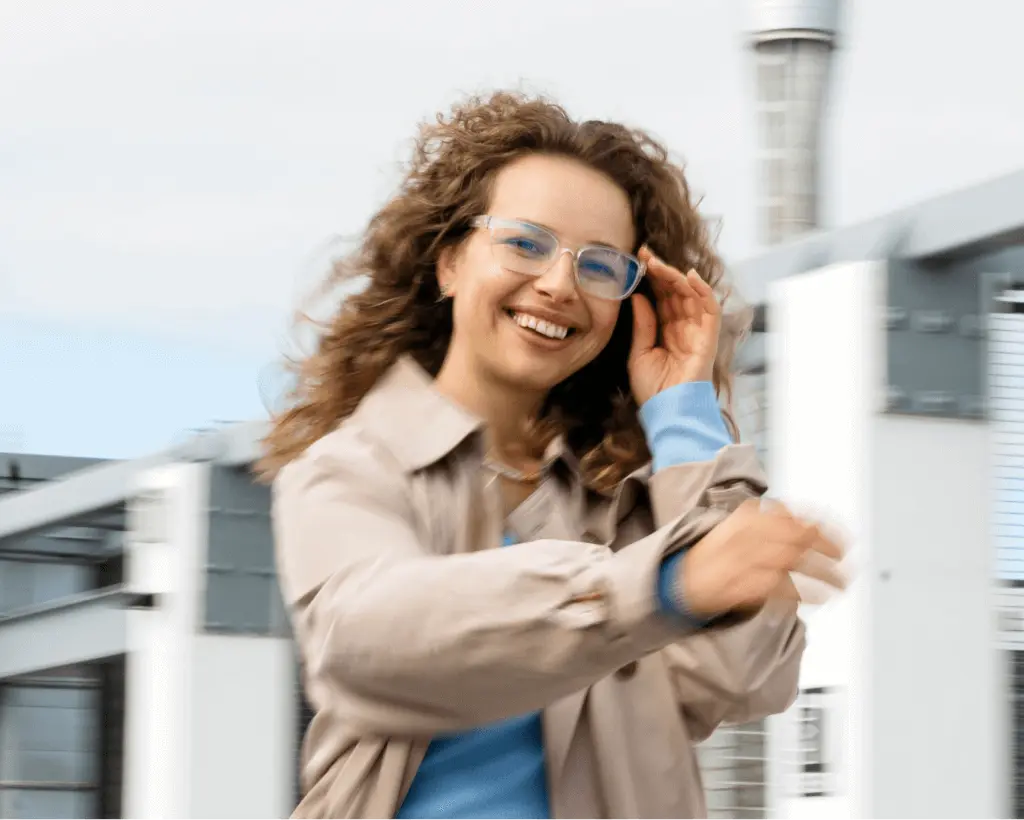 Dynamic portrait of a woman wearing a Dresden Vision glasses and a light coat, smiling while moving outdoors near a modern building