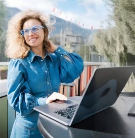 A smiling woman wearing stylish blue Dresden Vision Australia glasses works on her laptop outdoors, embracing comfort and clarity in sustainable eyewear.