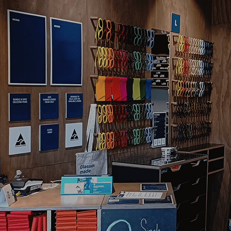 A colorful display of eyewear frames on wooden peg racks inside the Dresden Vision West End store, featuring plywood walls and a minimalist service counter.