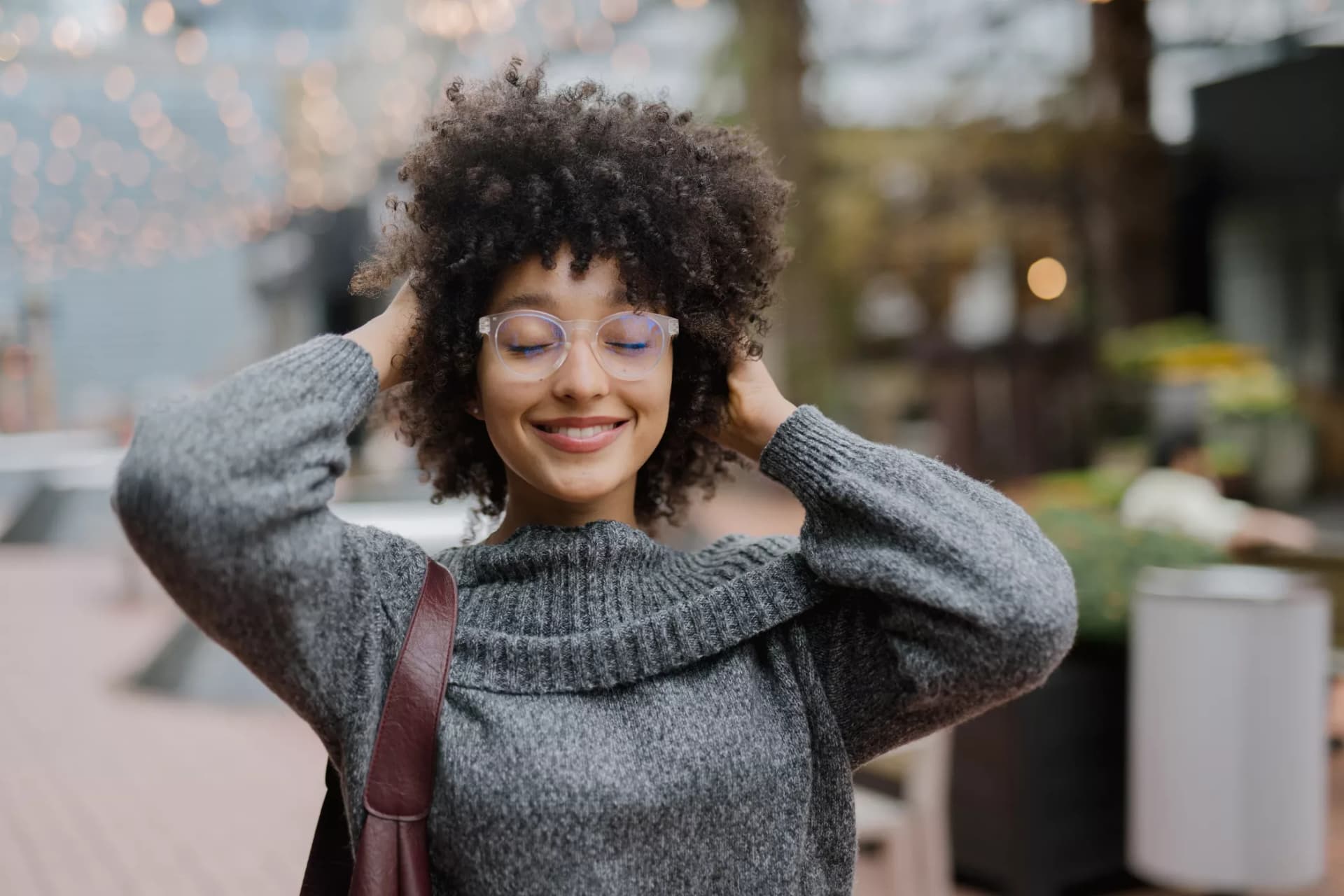 Smiling woman wearing bifocal lenses, grey sweater, and shoulder bag, standing outdoors with hands in curly hair.